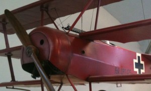 The image shows a close-up view of a red triplane, a type of vintage aircraft with three wings stacked vertically. The plane has a wooden propeller and a black cross insignia on its side, indicating it is likely a replica or model of a World War I-era German fighter plane. The aircraft is displayed indoors, possibly in a museum or exhibition setting.