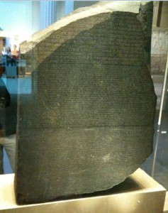 The image shows the Rosetta Stone, an ancient Egyptian artifact made of granodiorite. It is inscribed with a decree issued in Memphis, Egypt in 196 BC, written in three scripts: Greek, Demotic, and Hieroglyphic. The stone is displayed in a museum setting, encased in glass.