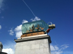 The image shows a large sculpture of a ship inside a glass bottle. The bottle is mounted on a stone pedestal against a backdrop of a clear blue sky with a few clouds. The ship inside the bottle appears to be detailed with sails and masts.