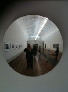 The image shows a reflection in a circular mirror mounted on a wall in an art gallery. The gallery has a high ceiling with skylights and wooden flooring. Several framed artworks are displayed on the walls. There are a few people visible in the reflection, including the person taking the photo.