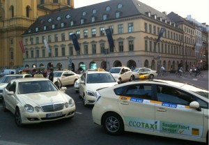 The image shows a busy street scene with several white taxis, including an "EcoTaxi," parked or driving in front of a large, historic building with a sloped roof and multiple windows. The building has several flags hanging from it, and there are other buildings visible in the background. The scene appears to be in an urban area, possibly in a European city.