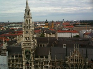 The image shows an aerial view of a cityscape with a prominent Gothic-style building in the foreground. The building features a tall clock tower with intricate architectural details. The city extends into the background with various other buildings, including a notable yellow structure with twin towers. The sky is overcast, adding a muted tone to the scene.