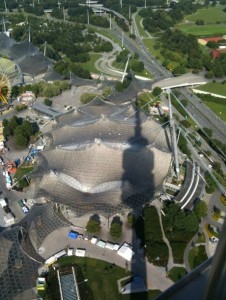 Aerial view of a large, modern architectural structure with a distinctive, tent-like roof design. The structure is surrounded by green spaces, roads, and pathways. The shadow of a tall tower or structure is cast over the building and the surrounding area. The scene includes various smaller buildings and vehicles, indicating an urban environment.