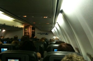 The image shows the interior of an airplane cabin. Passengers are seated, and the view is from the back of the cabin looking forward. The overhead compartments are closed, and individual screens are visible on the backs of the seats. The lighting is dim, suggesting it might be during a night flight or a time when the cabin lights are turned down.