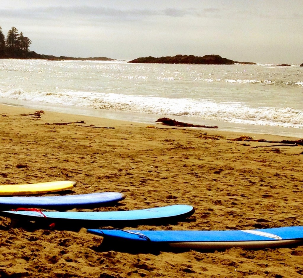 The image shows a sandy beach with several surfboards lying on the sand near the shoreline. The surfboards are in various colors, including blue and yellow. The ocean waves are gently crashing onto the shore, and there are some small islands or rocks visible in the distance. The sky is partly cloudy, and the overall scene appears calm and serene.