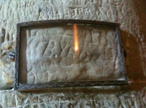 The image shows a stone surface with an inscription that appears to be protected by a rectangular glass frame with a decorative border. The inscription on the stone reads "B. Love" and is carved into the stone. The glass frame is secured to the stone surface with metal fasteners on either side. The stone surface around the inscription also has various other carvings and markings.