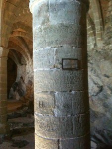 The image shows a stone column inside an ancient or historical structure. The column is cylindrical and made of large stone blocks stacked on top of each other. The surface of the column appears weathered and has some inscriptions or carvings on it. In the background, there are stone arches and walls, suggesting that the location might be a historical site or ruin. The lighting is dim, adding to the ancient atmosphere of the scene.