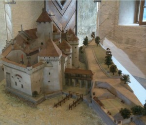 A detailed model of a medieval castle is displayed in the image. The model features various structures including towers, walls, and a gatehouse. The castle is surrounded by a moat and has a bridge leading to the entrance. There are also miniature trees and pathways around the castle, adding to the realistic representation. The model is set on a textured base that mimics the landscape.