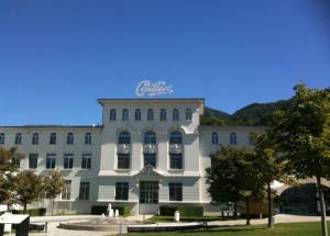 The image shows a large, white, multi-story building with a sign on the roof that reads "Cailler." The building has several arched windows and is surrounded by a well-maintained lawn with a few trees. The sky is clear and blue, and there are mountains visible in the background.