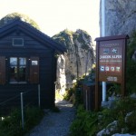 The image shows a small, dark wooden cabin with closed shutters on its windows, situated on a narrow gravel path. To the right of the cabin, there is a large sign that reads "La Rambertia Jardin Alpin" and provides additional information about the alpine garden, including its founding year (1896) and altitude (2000 meters). The sign also indicates free entry. The background features rocky cliffs and lush greenery, suggesting a mountainous location.