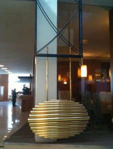 The image shows a modern indoor sculpture in a lobby area. The sculpture consists of horizontal, gold-colored, cylindrical bars stacked in a spherical shape. It is mounted on a base in front of a tall, white column. The background includes a seating area with people, warm lighting, and a reception desk. The overall setting appears to be a hotel or an office building lobby.