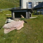 The image shows a grassy outdoor area with a small structure that appears to be an animal enclosure. There are several marmots visible in the scene. One marmot is sitting near the entrance of the enclosure, another is on the grass to the left, and a third is partially visible near some rocks in the foreground. The enclosure is situated near a building with a window and shutters, and there is another structure with a glass wall to the right. The area is surrounded by a fence and has a natural, hilly landscape.