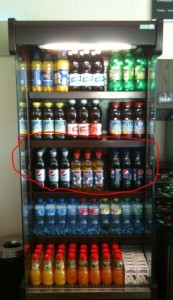 A glass-front refrigerator is stocked with various beverages. The top shelf contains bottles of yellow and green drinks. The second shelf has a mix of dark-colored bottles and cans. The third shelf, which is circled in red, contains bottles of Pepsi and other similar beverages. The fourth shelf has bottles of water. The bottom shelf is filled with bottles of orange and red drinks.