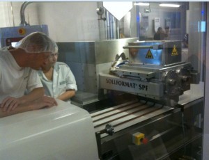 Two people wearing white lab coats and hairnets are observing a machine labeled "SOLIFORMAT SPF" in a factory or laboratory setting. The machine appears to be part of a production line, possibly for food or pharmaceutical products. The environment is clean and industrial, with stainless steel equipment and safety signs visible.