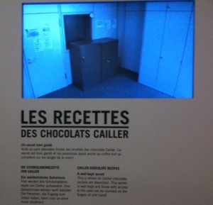 The image shows a display with text and a photograph. The text is in multiple languages, including French, German, and English. The French title reads "LES RECETTES DES CHOCOLATS CAILLER," which translates to "CAILLER CHOCOLATE RECIPES" in English. Below the title, there is a paragraph of text in each language. Above the text, there is a photograph of a room with a cabinet and some doors, all bathed in blue light. The display appears to be part of an exhibit, possibly in a museum or a chocolate factory tour.