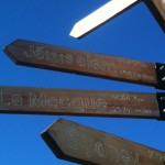 The image shows a wooden signpost with three directional arrows pointing to different cities. The top arrow points to "Jérusalem" with a distance of 2903 km (1807 miles). The middle arrow points to "La Mecque" with a distance of 4064 km (2524 miles). The bottom arrow points to "Sydney" with a distance of 16724 km. The sky in the background is clear and blue.