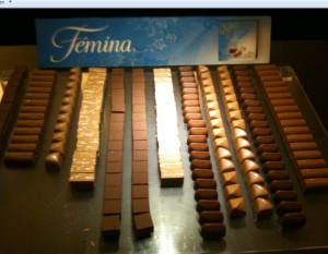 The image shows an assortment of chocolates arranged in neat rows on a reflective surface. There are different types of chocolates, including dark, milk, and white varieties, each with distinct shapes such as rectangular bars, squares, and rounded pieces. In the background, there is a blue and white sign with the word "Femina" written on it.