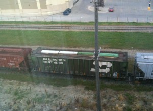 The image shows a train with several freight cars on a railway track. The train is viewed from above, and the freight car in the center has the text "BURLINGTON NORTHERN" written on it. There is a utility pole in the foreground, and a grassy area separates the railway tracks from a paved area and a building in the background.