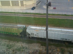 The image shows a view from above of two freight train cars on parallel tracks. One train car is green, and the other is white. In the background, there is a parking lot with a few cars and a large building. A utility pole is visible in the foreground, and there is some grass and vegetation near the train tracks.