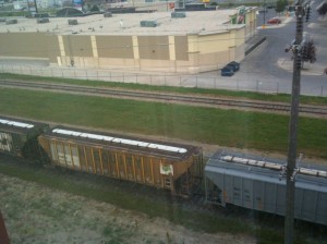 The image shows a view from a higher vantage point, looking down at a train with several freight cars on a railway track. The train is passing through an area with grass on either side of the tracks. In the background, there is a large commercial building with a parking lot and a few cars. A utility pole is also visible in the foreground.