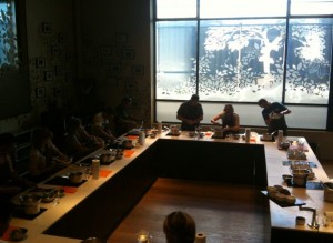 The image shows a group of people participating in a cooking class. They are seated around a large, U-shaped kitchen counter with various cooking utensils and ingredients in front of them. Three individuals are standing at the front, demonstrating cooking techniques. The room has a large window with decorative patterns, allowing natural light to illuminate the space. The atmosphere appears to be focused and educational.