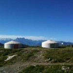 The image shows two traditional yurts (circular tents) set on a grassy hillside with a backdrop of distant mountains under a clear blue sky. The yurts have white coverings with one having a red door and the other a green door. The landscape is rugged with patches of grass and rocks. There is a person standing near the yurt on the right side of the image.