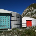 The image shows two traditional yurts (portable, round tents covered with skins or felt) set against a mountainous backdrop. The yurt in the foreground has a turquoise door with intricate designs, while the yurt in the background has a red door with similar decorative patterns. The sky is clear and blue, and the terrain around the yurts is rocky with patches of grass.