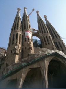 The image shows the Sagrada Família, a large unfinished Roman Catholic basilica in Barcelona, Spain. The structure features multiple tall, intricately designed spires and is under construction, with scaffolding visible around the lower sections. The sky is clear and blue in the background.