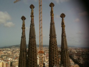 The image shows the spires of the Sagrada Família, a large unfinished Roman Catholic church in Barcelona, Spain. The spires are tall and intricately designed, with a construction crane visible in the background. The cityscape of Barcelona can be seen in the distance under a partly cloudy sky.