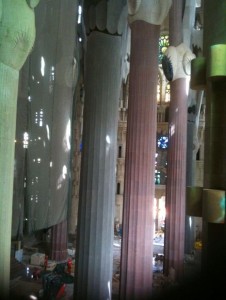 The image shows the interior of a large, ornate building with tall, intricately designed columns. The columns are of varying colors and textures, and some have decorative elements at the top. There is scaffolding and construction equipment visible at the base of the columns, indicating that the building is under construction or renovation. Light filters through the space, creating a pattern of shadows and highlights on the columns and the floor. The overall atmosphere is grand and architectural.