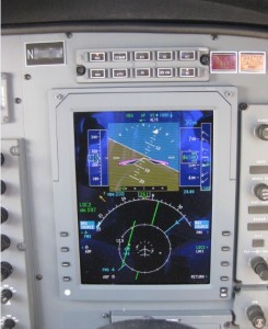 The image shows a close-up view of an aircraft's cockpit display panel. The central screen displays flight information, including altitude, speed, and navigation data. The screen is divided into sections, with a map and flight path on the top half and various instrument readings on the bottom half. Surrounding the screen are various buttons, switches, and knobs used for controlling the aircraft's systems.