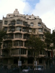 The image shows a unique, multi-story building with an undulating stone facade and irregularly shaped windows and balconies. The architectural style is distinctive, with organic, flowing lines and wrought iron balcony railings. There are trees in front of the building, and cars are parked along the street at the base of the building. The sky is partly cloudy.