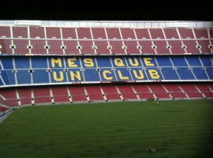 The image shows the interior of a large football stadium with red and blue seats. The seats in the middle section spell out the phrase "MÉS QUE UN CLUB" in yellow letters, which is Catalan for "More than a club." The field is green and appears to be well-maintained. There are no players or spectators visible in the image.