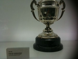 The image shows a silver trophy with two handles on either side, placed on a black base. There is a small plaque or label next to the trophy, but the text on it is not fully legible. The background is plain and white.