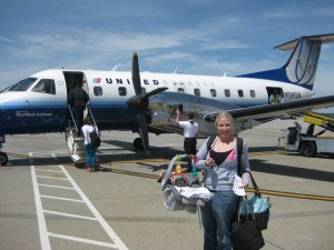 A woman is standing on an airport tarmac in front of a small United Express airplane. She is holding a baby carrier and a bag. In the background, other passengers are boarding the plane via a staircase. The sky is clear and blue.