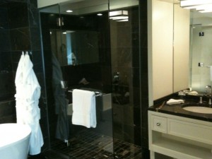 The image shows a modern bathroom with a glass-enclosed shower area. There is a white bathrobe hanging on the wall next to the shower. A white towel is draped over the shower door. The bathroom has a sink with a countertop and a mirror above it. The overall design is sleek and contemporary, with dark tiles and light-colored fixtures.