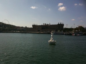 The image shows a body of water with a white statue of a person standing on a small platform in the foreground. In the background, there is a large, historic-looking building with multiple spires and ornate architecture. The sky is clear with a few scattered clouds, and there are some boats docked near the building. The scene appears to be taken during the daytime.