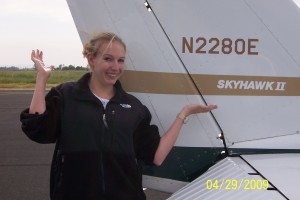 A person is standing next to the tail of an airplane, smiling and gesturing with both hands. The tail of the airplane has the registration number "N2280E" and the model name "SKYHAWK II" written on it. The person is wearing a black jacket and has blonde hair. The date "04/29/2009" is visible in yellow text at the bottom right corner of the image.