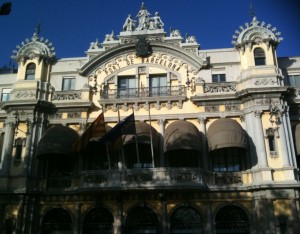 The image shows an ornate, historic building with intricate architectural details. The facade features multiple arched windows, decorative columns, and elaborate sculptures. There are three flags displayed in front of the building, and the structure is bathed in sunlight against a clear blue sky. The building has a grand and majestic appearance, with a combination of classical and baroque elements.