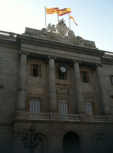 The image shows the facade of a historic building with classical architectural elements, including tall columns and a decorative crest at the top. Two flags are flying above the building, one of which appears to be the flag of Spain. There is a clock situated between the columns on the upper part of the building. The structure has large windows and a balcony with a balustrade. The sky is clear, indicating it is daytime.