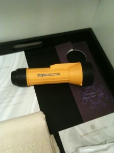 The image shows a yellow flashlight with black ends placed on a dark surface. The flashlight has the brand name "Eveready" printed on it. Surrounding the flashlight are various papers and documents, including a white sheet and a purple card. There is also a beige cloth partially visible in the bottom left corner.