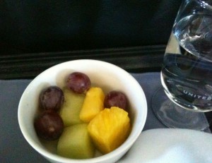 A small white bowl containing a mix of fruit pieces, including red grapes, chunks of pineapple, and pieces of melon. Next to the bowl is a glass of water with the word "United" printed on it. The items are placed on a tray with a dark background.