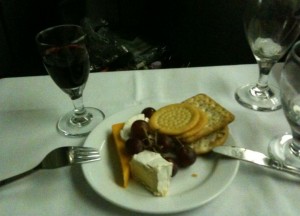 A plate with a selection of cheeses, crackers, and grapes is placed on a white tablecloth. There is a fork to the left of the plate and a knife to the right. A glass of red wine is positioned to the left of the plate, and an empty wine glass is to the right.