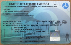 The image is of a United States of America Federal Aviation Administration (FAA) pilot certificate. The certificate includes the following details:

- Title: "UNITED STATES OF AMERICA"
- Issuing Authority: "DEPARTMENT OF TRANSPORTATION - FEDERAL AVIATION ADMINISTRATION"
- Name: "MICHELLE [REDACTED] SINGH"
- Address: [REDACTED]
- Nationality: "USA"
- Date of Birth: "10 FEB"
- Sex: "F"
- Height: "67"
- Weight: "140"
- Hair: "BLOND"
- Eyes: "GREEN"
- Certificate Type: "PRIVATE PILOT"
- Certificate Number: [REDACTED]
- Date of Issue: "29 APR 2009"
- Signature of the Administrator
- A holographic seal and a background image of an aircraft and a pilot.

(Note: Personal information has been redacted for privacy.)