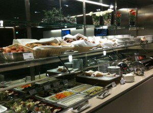 The image shows a buffet setup with various food items displayed in trays and containers. The buffet includes a variety of dishes such as salads, vegetables, and possibly meats or pastries. There are also plates and utensils available for serving. Above the food, there are decorative plants and flowers. The setting appears to be well-lit and organized, likely in a restaurant or dining area.