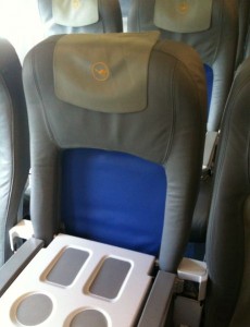 The image shows a row of airplane seats. The seats are upholstered in dark gray and blue fabric. The headrests have a logo on them, and the middle seat has its tray table down, revealing cup holders. The seats are arranged in a typical economy class configuration.