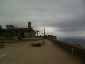 The image shows a cloudy, overcast day at a historical fort or castle. The structure is made of stone, with visible battlements and a cannon pointing outwards. There are some benches and a pathway leading towards the edge of the fort, which overlooks a body of water in the distance. The overall atmosphere is moody and somewhat desolate.