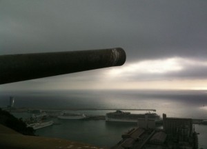 A large cannon is positioned in the foreground, pointing towards the horizon over a body of water. Below, there is a harbor with several docked ships and industrial buildings. The sky is overcast with a mix of dark clouds and a break of light near the horizon.