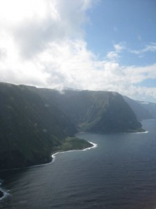 The image shows a coastal landscape with steep, green cliffs meeting the ocean. The sky is partly cloudy, and the sunlight is diffused through the clouds, casting a soft light on the scene. The water appears calm, and the coastline curves gently along the base of the cliffs.