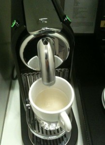 The image shows a coffee machine with a white ceramic mug placed under the dispenser. The machine appears to be in the process of brewing coffee, as there is a light-colored liquid in the mug. The coffee machine has a sleek, modern design with a metallic finish.